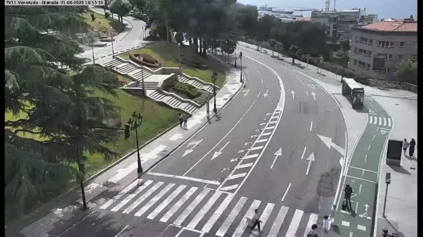 An aerial view shows a curving road with a crosswalk and bike lane, flanked by greenery, stairs, and buildings, on a partly sunny day.