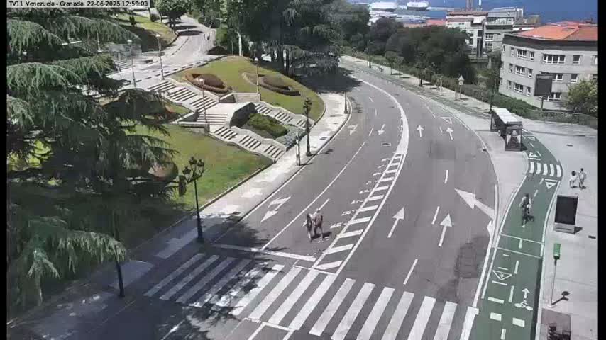 A mostly empty roadway curves through a park-like setting on a sunny day, with pedestrians and a cyclist visible, near a building.