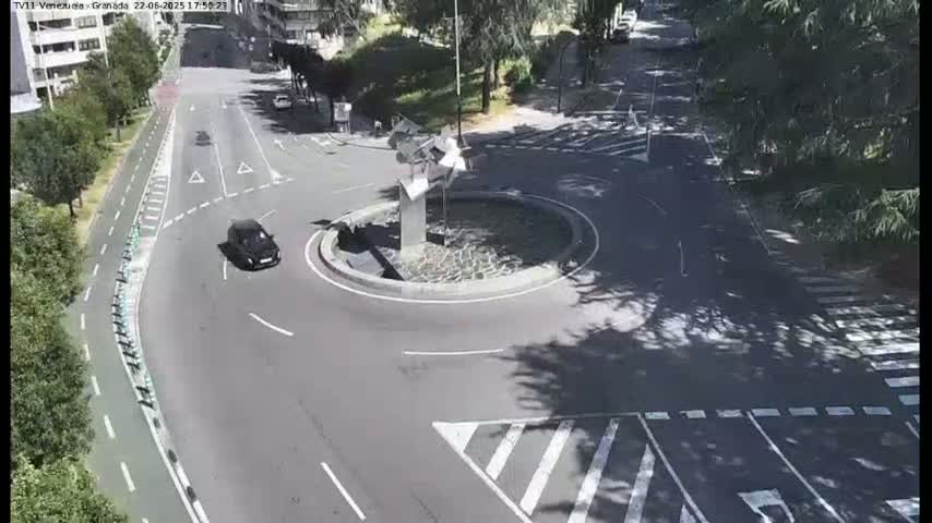 A dark-colored car drives around a roundabout with a modern sculpture in the center, under a sunny sky.