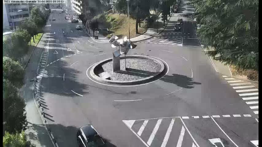 A dark-colored car approaches a roundabout featuring a metallic sculpture in the center, under a sunny sky.