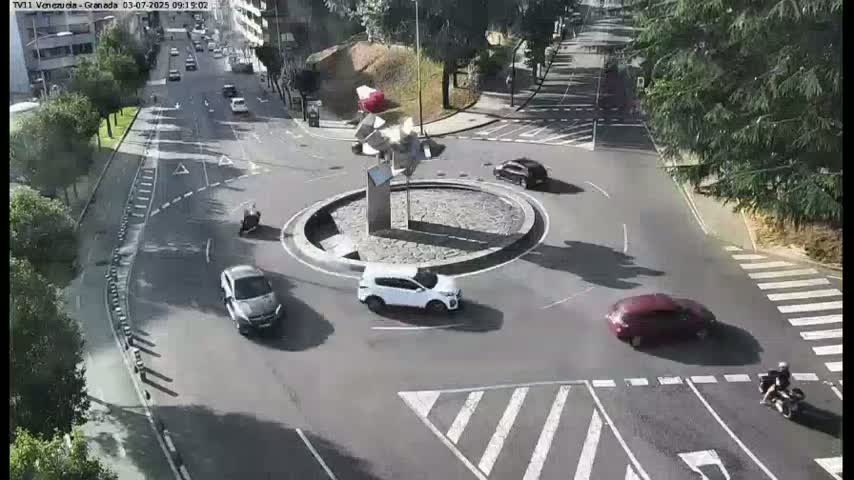 A sunny day shows several cars navigating a roundabout in the middle of a city street, with a modern sculpture in the center and a motorcyclist approaching.