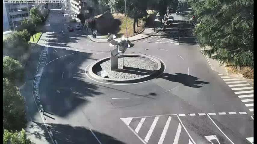 A sunny overhead view of a circular traffic island with a modern sculpture in the center, surrounded by roads with crosswalks and a few parked cars.