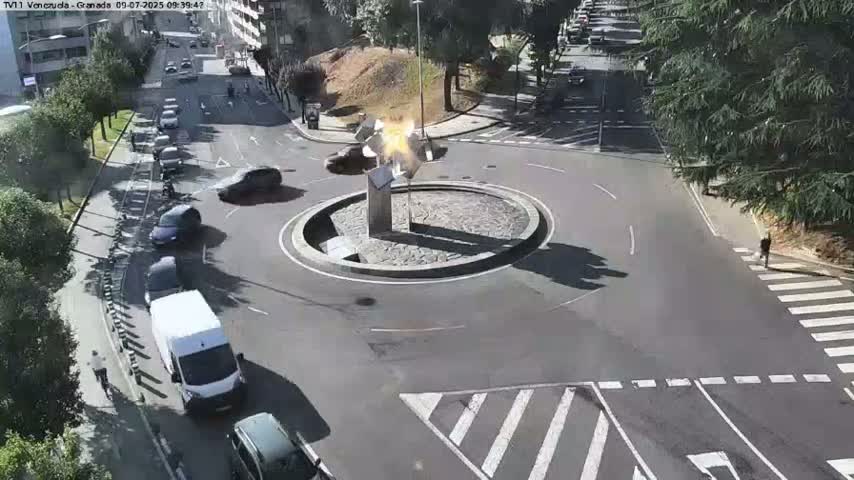 A traffic circle with a central sculpture is surrounded by cars and pedestrians on a sunny day.