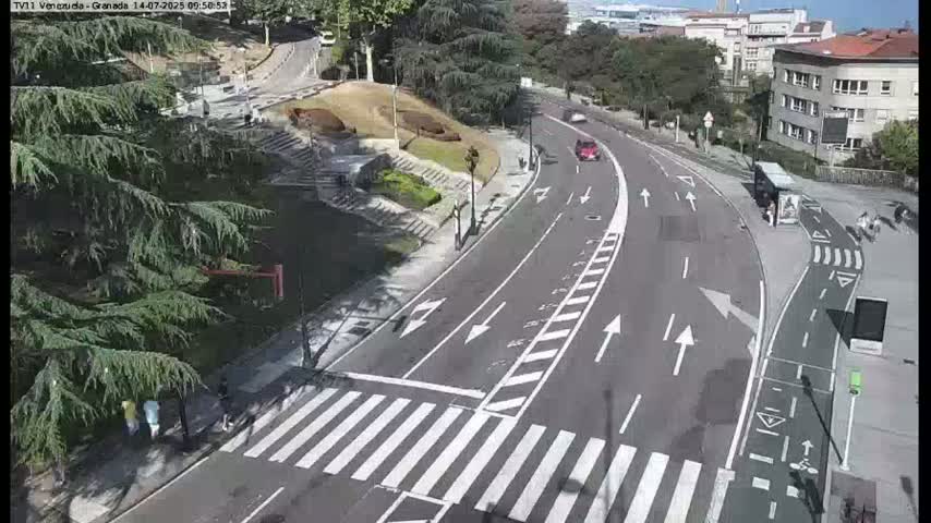 A curving road with a crosswalk and bike lane winds through a city, passing by green spaces and buildings under a sunny sky.