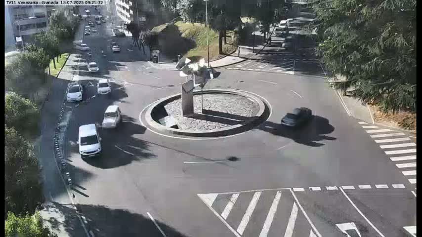 Several cars are driving around a roundabout with a modern sculpture in the center, on a sunny day.