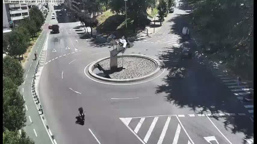 Several cars are driving around a roundabout with a modern sculpture in the center, on a sunny day.