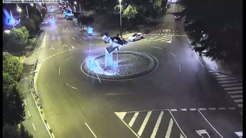 A nighttime aerial view shows a roundabout with a modern sculpture in the center, surrounded by roads with cars and pedestrian crossings.