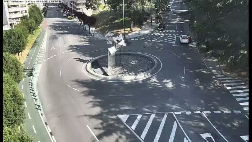 A nighttime aerial view shows a roundabout with a modern sculpture in the center, surrounded by roads with cars and pedestrian crossings.