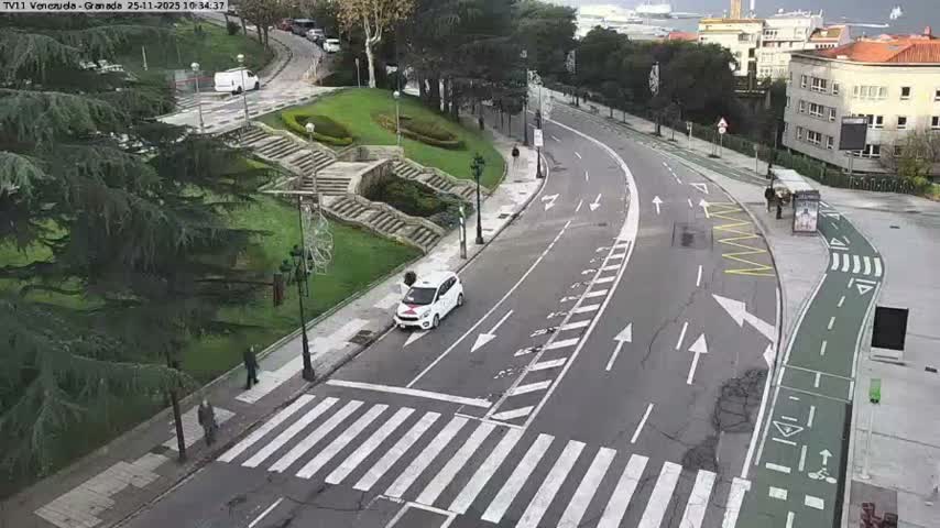 An aerial view captures a curved urban street featuring a multi-lane road, a dedicated green bike lane, and a crosswalk where a white car is stopped, with pedestrians on the sidewalks, lush green parkland with staircases to the left, and buildings leading to a harbor with ships in the distance on the right, all under an overcast sky.