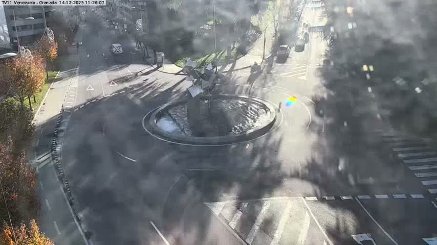 An aerial view captures a curved urban street featuring a multi-lane road, a dedicated green bike lane, and a crosswalk where a white car is stopped, with pedestrians on the sidewalks, lush green parkland with staircases to the left, and buildings leading to a harbor with ships in the distance on the right, all under an overcast sky.