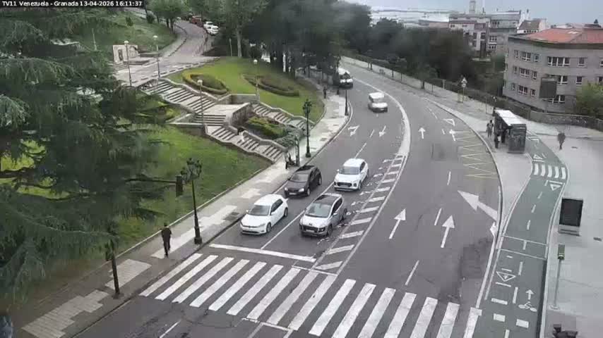 An aerial view captures a curved urban street featuring a multi-lane road, a dedicated green bike lane, and a crosswalk where a white car is stopped, with pedestrians on the sidewalks, lush green parkland with staircases to the left, and buildings leading to a harbor with ships in the distance on the right, all under an overcast sky.