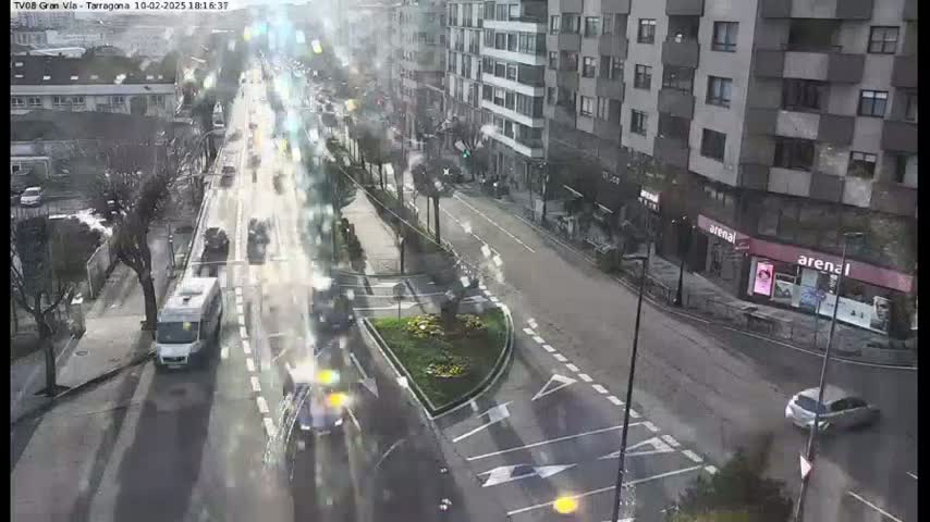 A city street scene with moderate traffic on a somewhat overcast day shows vehicles moving along a multi-lane road that passes a small traffic circle and several buildings.