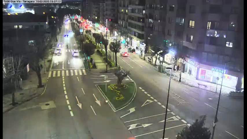 A nighttime street scene shows several cars driving along a multi-lane road that intersects with a smaller road, flanked by buildings and trees.