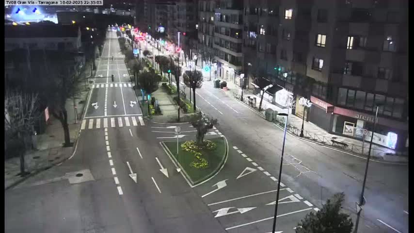 A nighttime, high-angle view shows a mostly empty, multi-lane road intersecting with a smaller road, bordered by buildings and lined with trees and streetlights.