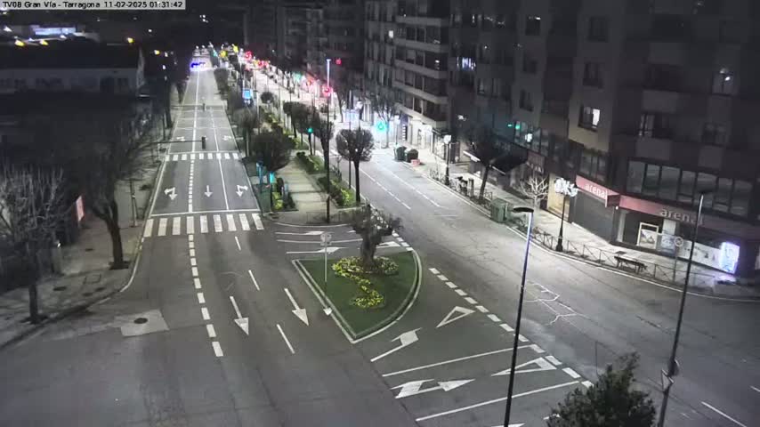 A mostly empty, well-lit city street at night, with buildings lining both sides and a small traffic circle in the center.