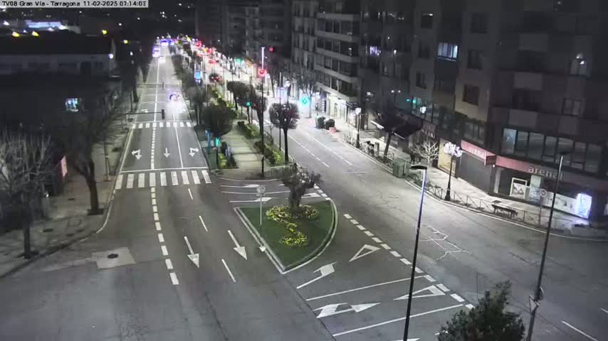 A mostly empty, well-lit city street at night, with buildings lining both sides and a small traffic circle in the center.