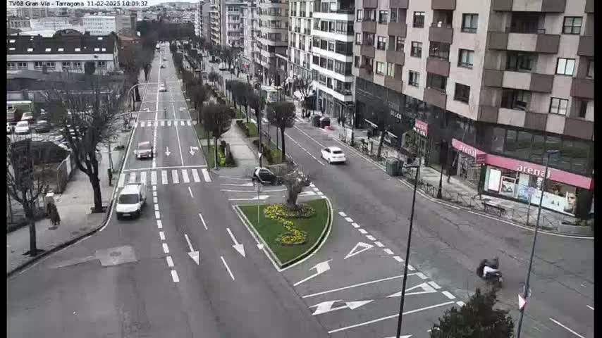 A street intersection with several cars and pedestrians, bordered by buildings, with a small landscaped area in the center under a clear sky.