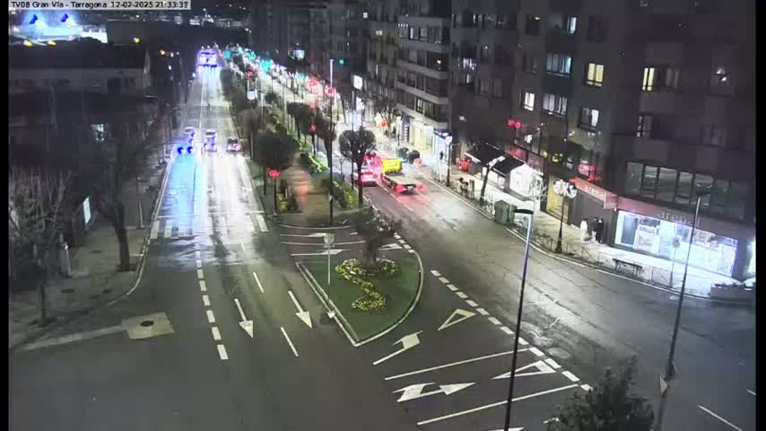A wet, nighttime street scene shows a multi-lane road with sparse traffic, bordered by buildings and trees.