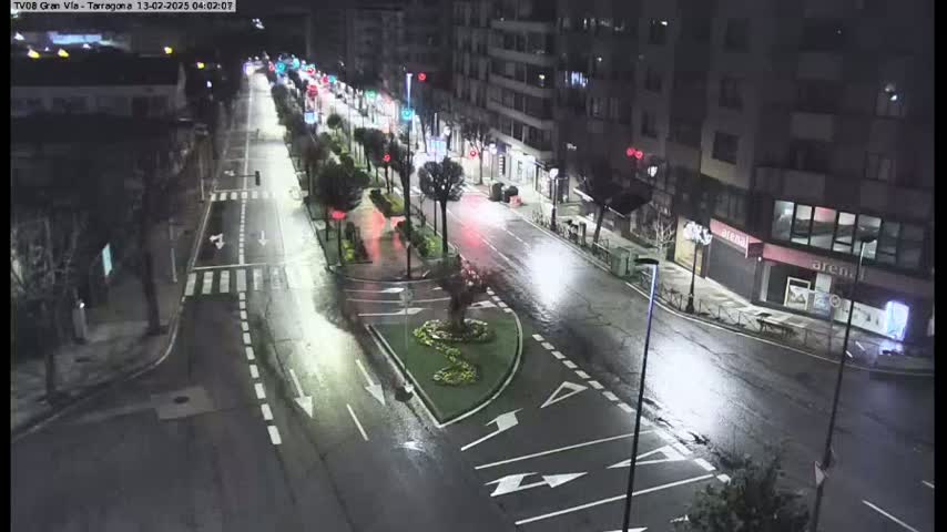 A wet, empty street at night is lit by streetlights, with buildings lining both sides and sparse landscaping in the center.