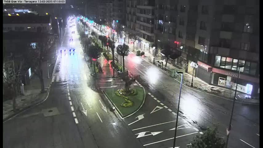 A wet, nighttime street scene shows several cars driving on a multi-lane road, lined with buildings and trees, under streetlights.