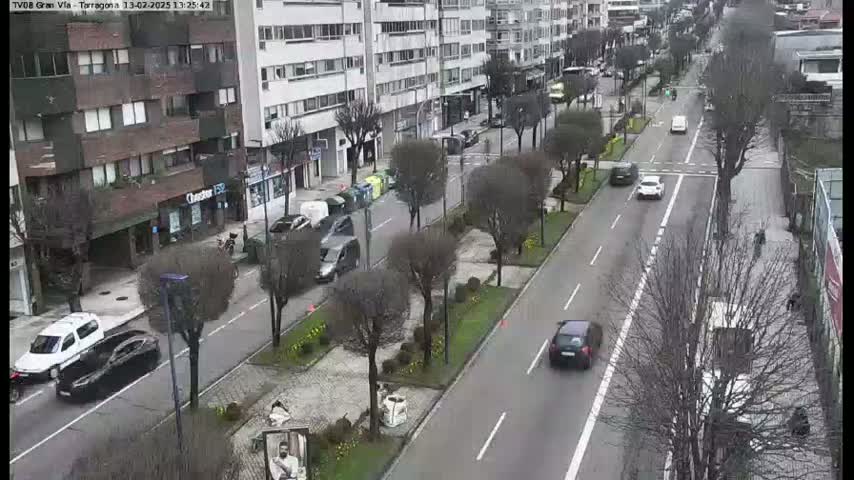 A city street with several cars driving on it, lined with buildings and bare trees under an overcast sky.