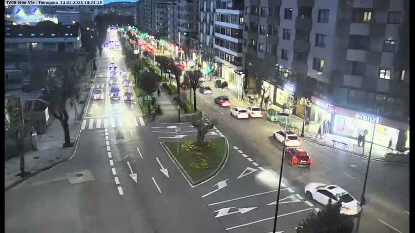 A nighttime, city street scene shows several cars driving along a multi-lane road that intersects with a smaller road, with buildings lining the street and a small landscaped median separating the lanes.