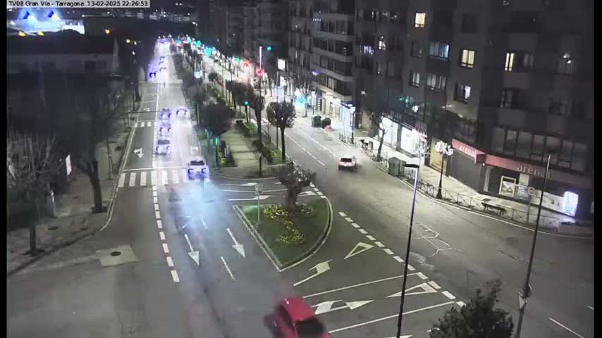 A nighttime street scene shows several vehicles traveling on a multi-lane road that intersects with another road, surrounded by buildings and streetlights.
