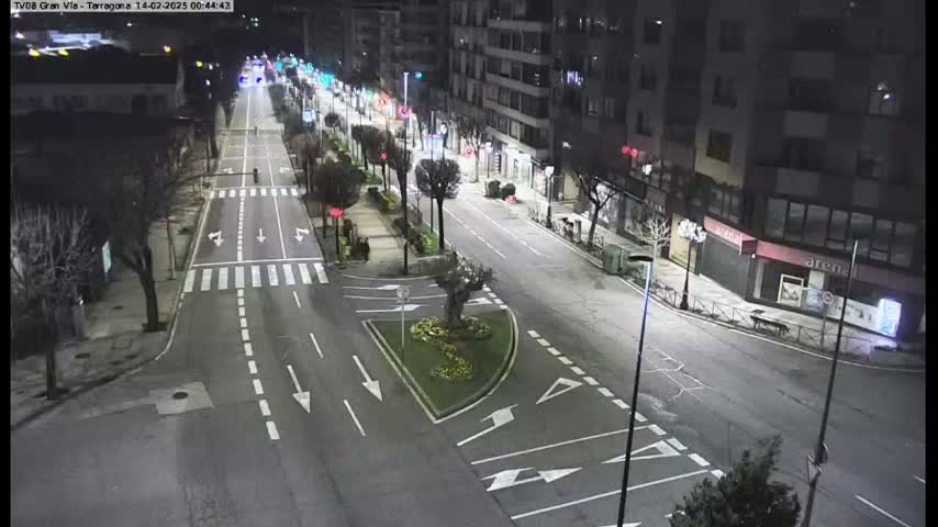 A mostly empty, well-lit city street at night, with a few cars visible in the distance.