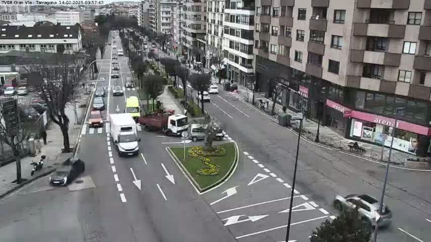 A city street scene on a partly cloudy day shows several cars and trucks navigating a multi-lane road with a landscaped traffic circle in the center.