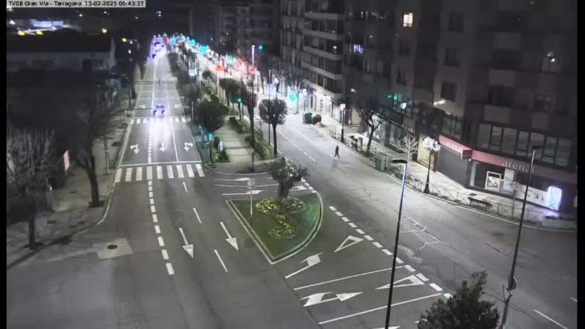 A mostly empty, well-lit city street at night, with a few cars driving on it and some pedestrians walking.