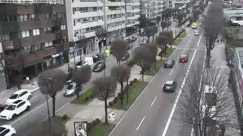 A city street scene on an overcast day shows several cars driving in both directions along a multi-lane road lined with trees and buildings.