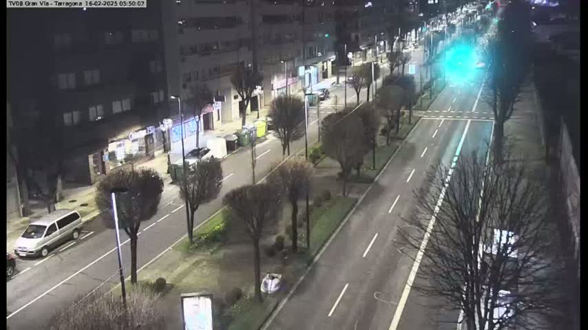 A nighttime, high-angle view of a city street with parked cars and sparse traffic, lined with trees and buildings.