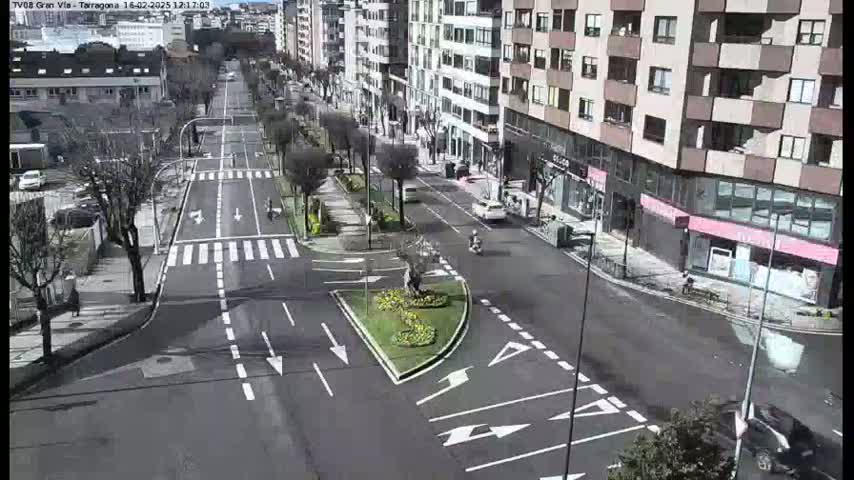 A sunny city street scene shows a multi-lane road intersecting with another road, surrounded by buildings and some landscaping.