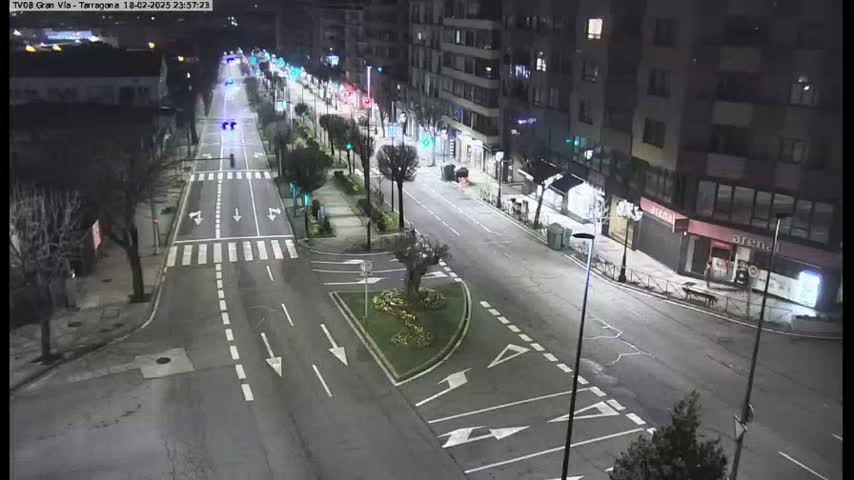 A nighttime, high-angle view of a mostly empty city street with buildings lining both sides, illuminated by streetlights, and a small median with landscaping in the center.