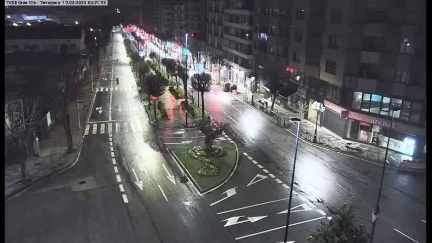 A wet, nighttime street scene shows a multi-lane road with sparse traffic, lined with buildings and trees, and a small, landscaped traffic island.