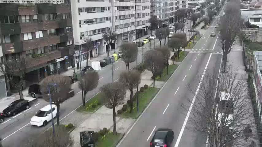 A mostly empty city street with parked and moving cars, lined with buildings and bare trees under a cloudy sky.