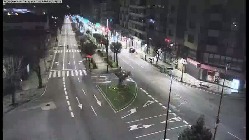 A nighttime, brightly lit street scene shows a multi-lane road with parked cars, buildings lining both sides of the road, and a small landscaped area in the center of the intersection.