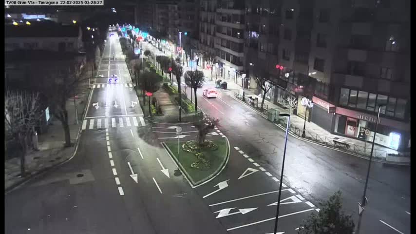 A nighttime, brightly lit street scene shows a sparsely trafficked road intersecting with a smaller road, lined with buildings and trees.