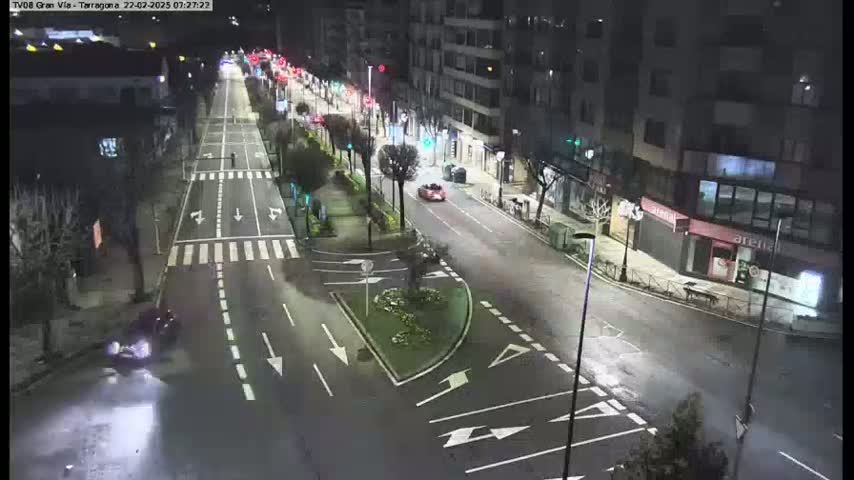 A wet, nighttime street scene shows a multi-lane road with sparse traffic, lined with buildings and trees, and featuring a small, landscaped median.
