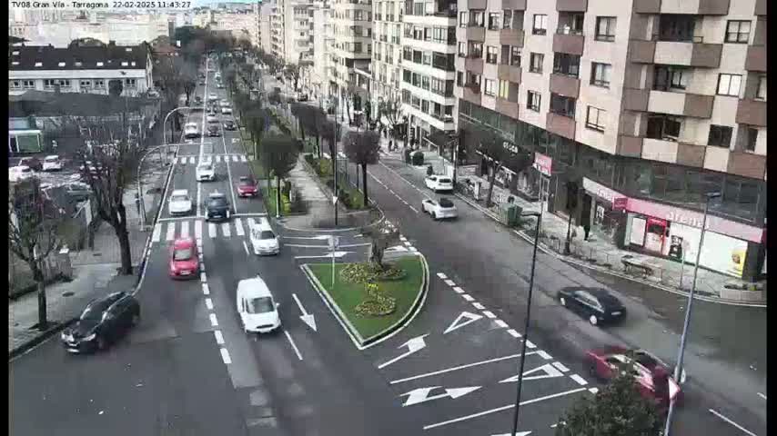 A high-angle view of a city street with moderate traffic on a partly cloudy day, featuring several cars navigating a roundabout and intersections.