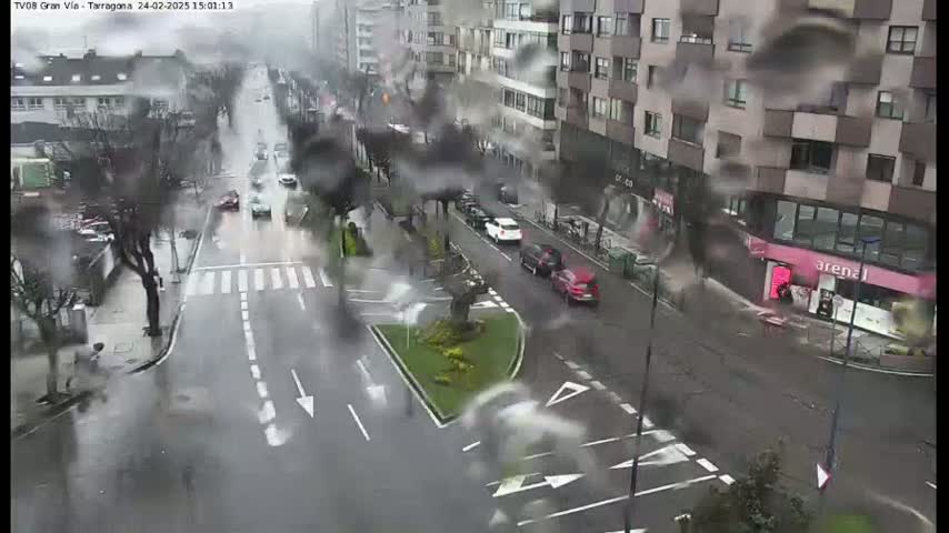 A rainy city street scene shows several cars driving on a wet road past buildings, some of which have shops on the ground floor.
