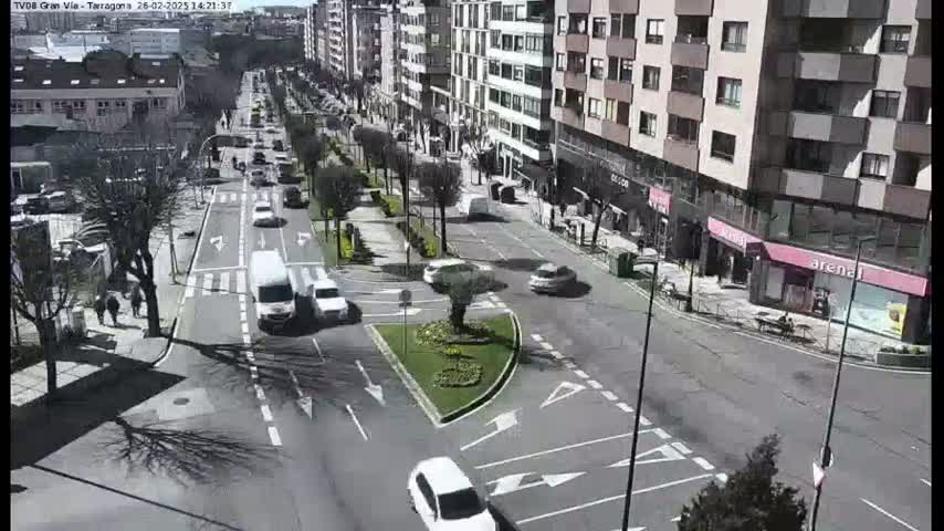 A sunny city street scene shows several cars driving on a multi-lane road, passing by buildings and trees.