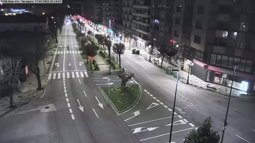 An empty, well-lit city street at night with buildings lining both sides, featuring a small traffic circle and sparse vegetation.