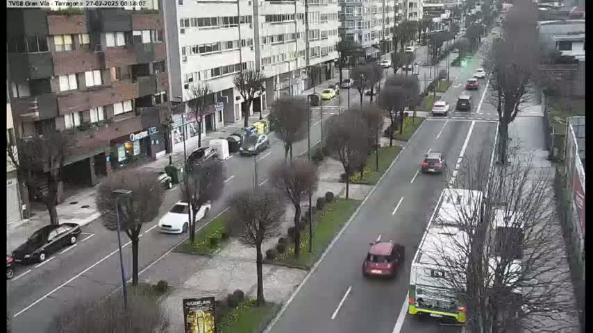 A city street scene on an overcast day shows several cars driving in both directions on a multi-lane road lined with trees and buildings.