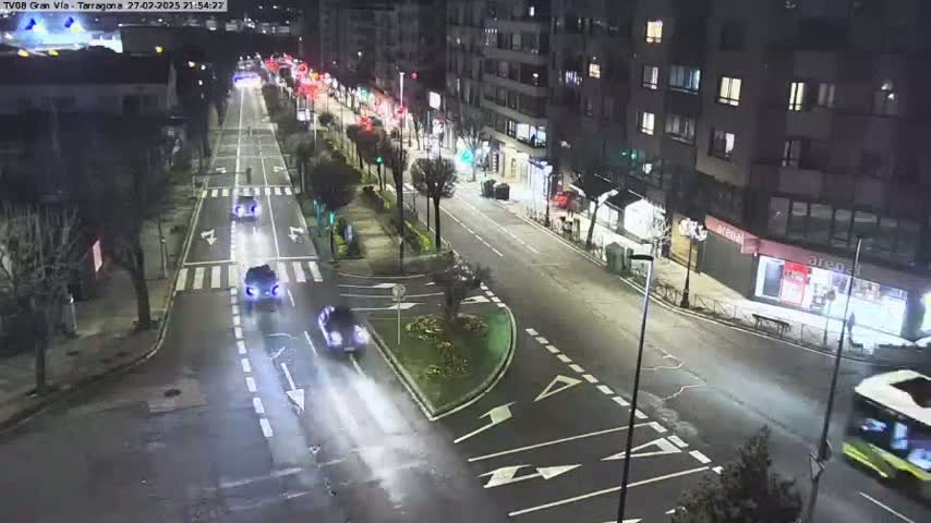 A nighttime view of a city street with several cars driving on a multi-lane road, lined with buildings and trees.