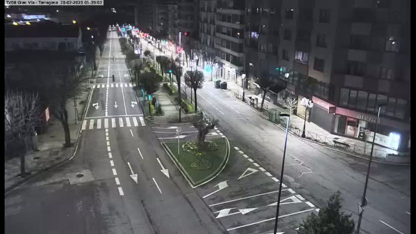 A mostly empty, city street at night is illuminated by streetlights, with sparse traffic and a small central garden visible.