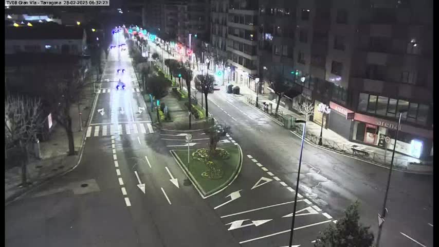 A wet, nighttime street scene shows a sparsely trafficked road with buildings lining both sides, streetlights illuminating the area.