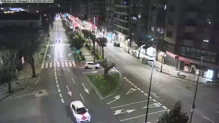 A nighttime, city street scene shows several cars driving on a well-lit road that intersects with another road, with buildings lining the street on both sides.