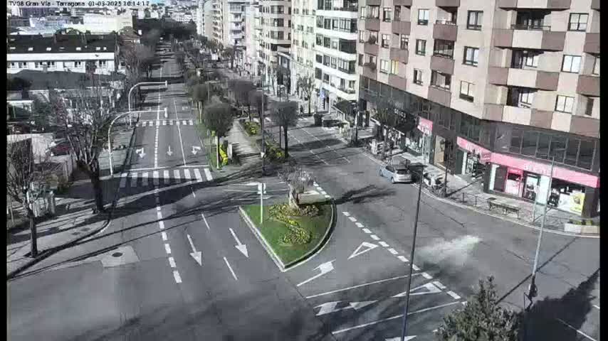 A sunny city street intersection shows sparse traffic, buildings lining the sides, and a small, landscaped median.