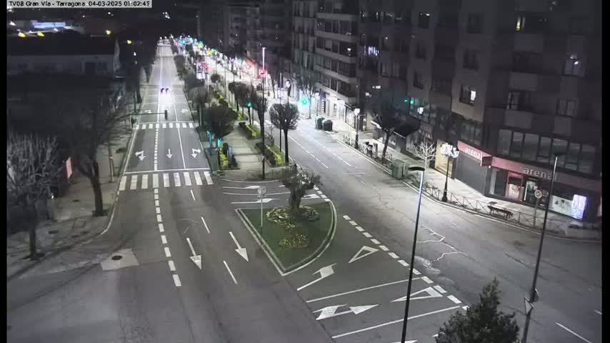 A mostly empty, well-lit city street at night shows several cars and pedestrians, with buildings lining both sides of the road.